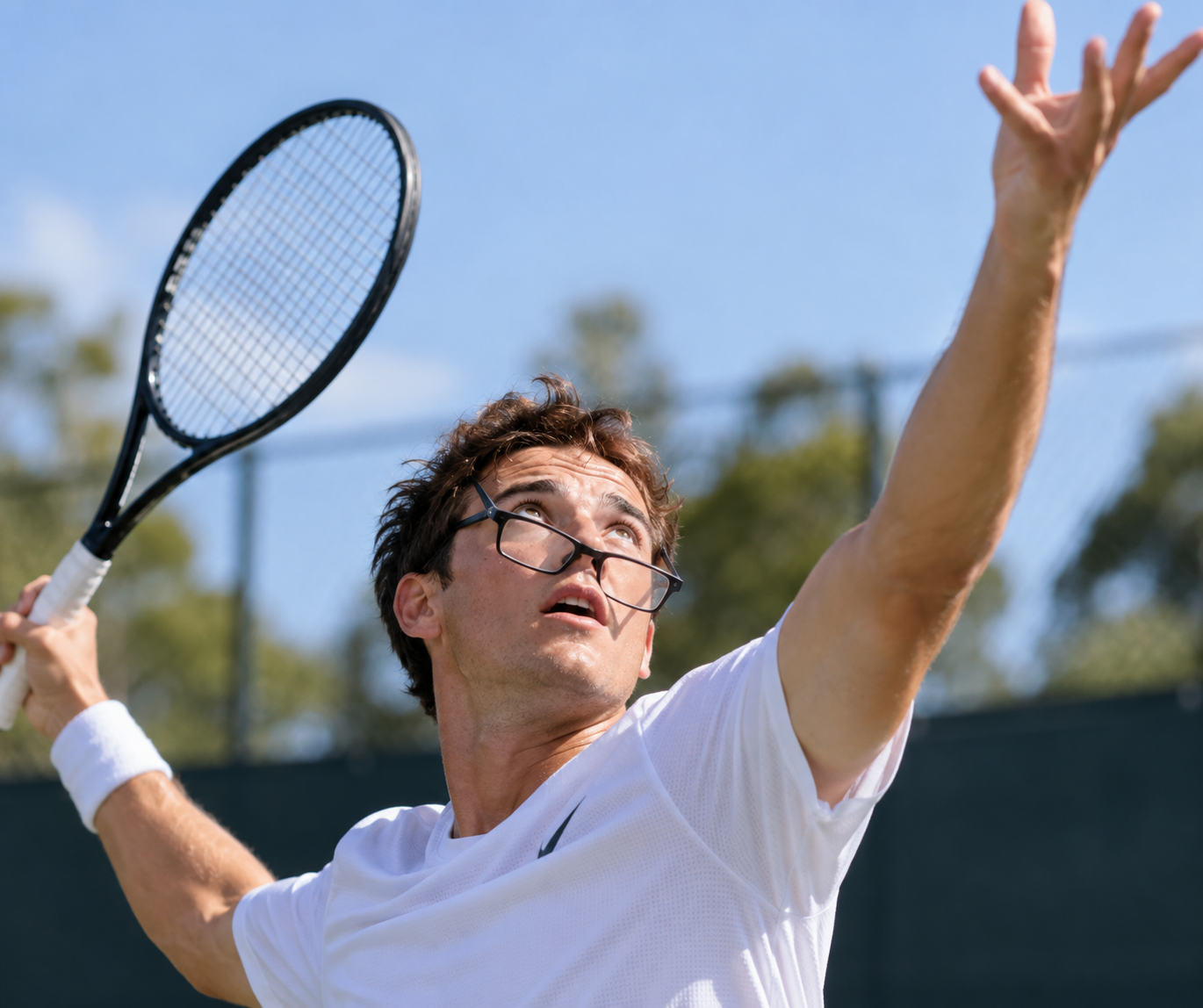 Tennis player serving while prescription glasses slip down during the ball toss, illustrating a common practical challenge for players considering personalised vision correction.